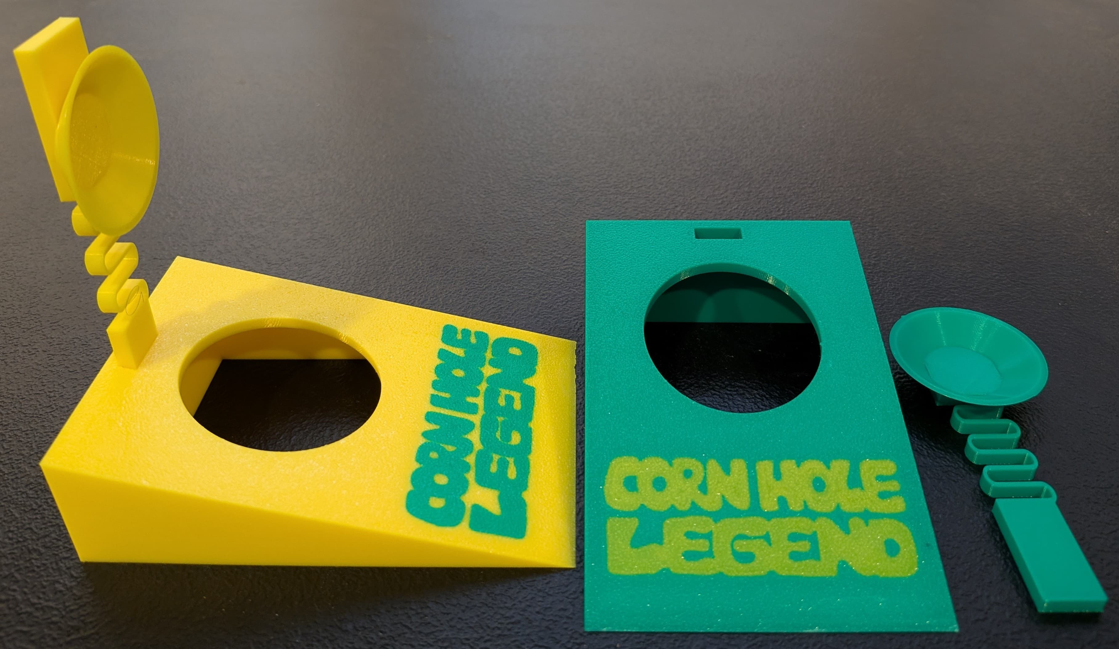 A close-up shot of three small, brightly colored 3D-printed objects on a dark surface. On the left is a green, angled stand with a circular opening and a green scoop attached by a flexible, coiled extension. In the center is a yellow rectangular piece with a circular cutout and the green text 'CORN HOLE LEGEND' printed on it. To the right is a separate yellow scoop with a flexible, coiled handle. The objects appear to be miniature game pieces or accessories.